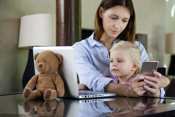 Cute little toddler girl sitting in front of laptop while mother is busy with her phone