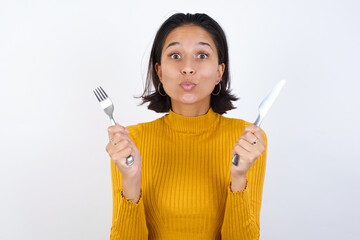 Young hispanic girl with short hair wearing casual yellow sweater isolated over white background hungry girl holding in hand fork knife want tasty yummy pizza pie