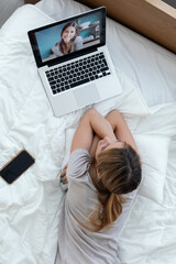 Beautiful young woman having videocall with laptop while explain something to her friend lying on the bed at home.