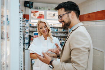 Obraz premium Young business man choosing and buying drugs in a drugstore while talking with attractive female pharmacist. She helping him with expert advices.