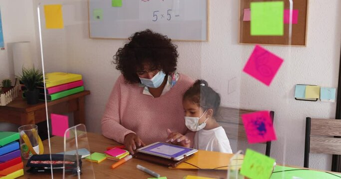 Teacher using tablet with child while wearing protective masks in preschool during coronavirus outbreak - Social distance at school concept