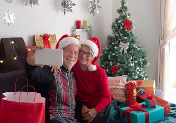 Obraz na płótnie Canvas Senior smiling couple of two grandparents with Santa Claus caps making selfie with Christmas tree and presents for the family on background. Love and family concept