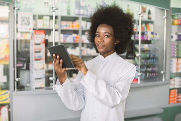 Pharmacy and healthcare concept, inventory. Young attractive smiling African American woman pharmacist or chemist writing on tablet while standing in modern pharmacy