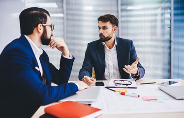 Confident male executive managers 30s discussing business strategy during working process at table desktop, Caucasian investors in formal wear have collaborative brainstorming in office interior