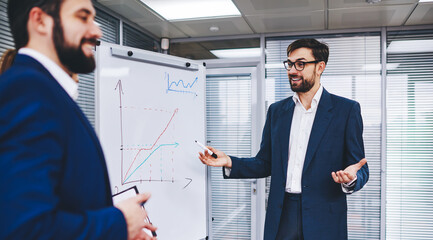 Group of successful financial experts enjoying friendly brainstorming for analyzing revenue infographics on flipchart, happy crew of experienced colleagues discussing diagram charts and smiling