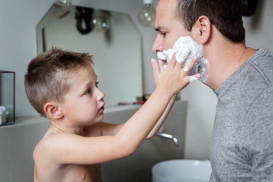 Young boy applying shaving cream on father's beard in bathroom at home
