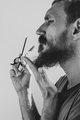 Fototapeta premium Adult man cutting his own beard and mustache with scissors and comb. Caucasian red bearded male trimming hair on face at home. Do it yourself self-made haircut.Selfcare DIY during quarantine isolation
