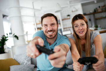  Husband and wife playing video game with joysticks in living room. Loving couple are playing video games at home