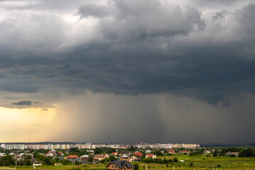 Moody landscape with dark stormy clouds with falling heavy downpour shower rain over distant town...