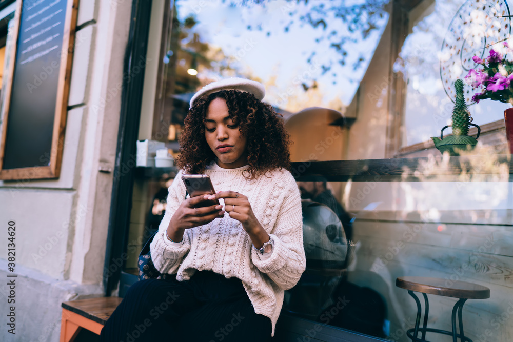 Poster pensive dark skinned curly woman using smartphone for reading messages and checking notification on 