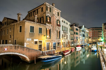Evening view of illuminated old buildings, bridges, floating boats and light reflections in canals water in Venice, Italy.