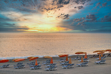Natural beach sunset landscape with multiple beach chairs and umbrellas standing on a fresh plowed stone sand in front of dramatic sunset.