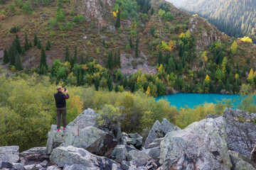 person on the edge of the mountain looking at the turquoise mountain lake surrounded by autumn colorful forest making photo using smartphone