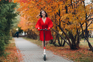 Young woman with electric scooter in red dress at the autumn city park