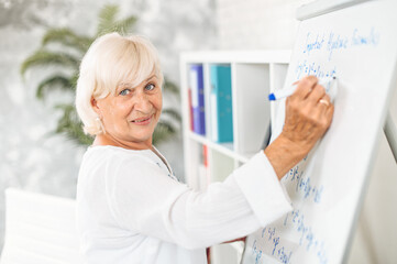 A senior woman in smart casual shirt is writing on flipchart and smiles. Online teacher, tutor....