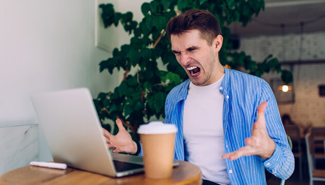 Angry stressed male freelancer irritated with bad internet connection sitting at desktop with netbook, unhappy emotional man 20s raging about software failure while downloading files on laptop