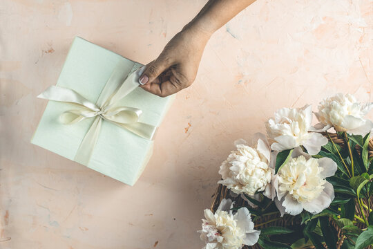 Womans hands hold ribbon for gift on light concrete table surface surrounded beautiful white peony flowers. Top view, flat lay, greeting card.