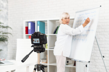 Elderly intelligent woman is writing on flipchart and recorded video classes on the camera, side view. Online teacher, video learning concept