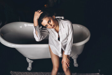 Top view of a young brunette with short hair sitting on the edge of a bath in a white shirt.