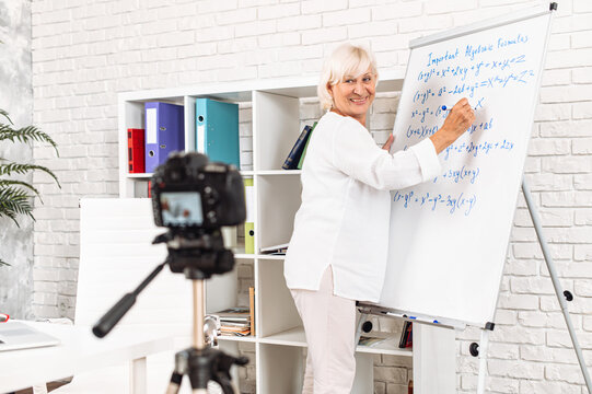 Cheerful senior female teacher writes algebra formulas on a whiteboard in modern classroom and records it on video. Online classes, online studying concept