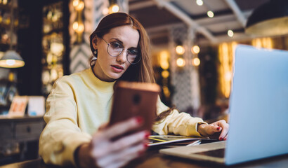 Caucasian hipster girl in spectacles reading received notification for cellphone application, millennial woman in classic eyewear using modern device during time for studying in coworking space