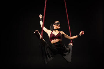 Brunette beautiful caucasian woman in Lace mask doing circus tricks on red silks isolated on black background.