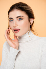 young woman in turtleneck looking at camera while touching face isolated on peach