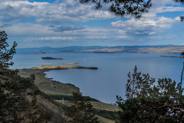 Green coniferous branches of pine trees on slope of mountain on coast of bay blue Baikal lake with islands and peninsula. Mountains on horizon. Sky with clouds. Siberia nature landscape