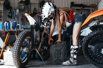 Plakat Female wearing shorts, motorcycle boots and cross-country helmet pouring gasoline into enduro motorcycle in the garage