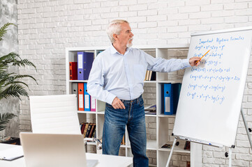Elderly teacher conducts an online lesson. Senior gray haired man with flipchart in front of laptop...