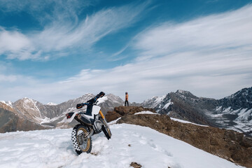 Female motorcyclist standing with her enduro motorcycle on snowy mountain top, snow peaks skyline...