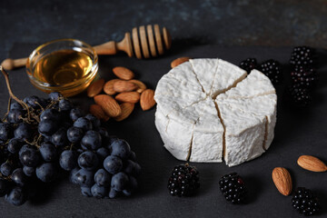 Camembert or brie french soft cheese with grapes, honey, nuts and berries on black stone serving board.