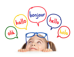 Beautiful cute little girl with eyeglasses hiding under table and looking at hello word in french, english, spanish, portuguese and german in speech balloons. K-12 foreign language learning concept.