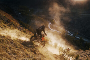 Extreme Rider climbing sand mountain top on off-road cross enduro motorcycle. Beautiful mountains landscape down on background, colourful autumn forest and river in sunshine - obrazy, fototapety, plakaty