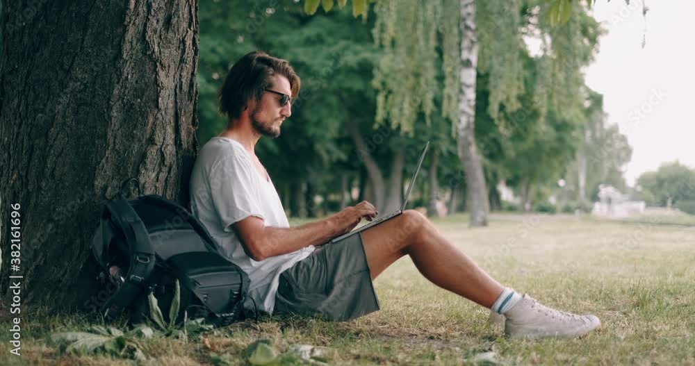 Poster bearded man is working with laptop at the park under the tree. happy freelancer is sitting and using