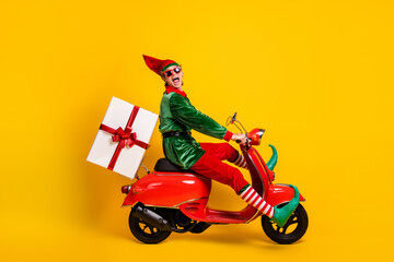 Profile side view of his he nice attractive cheerful cheery crazy funny guy elf riding moped delivering giftbox Eve Noel fairy isolated over bright vivid shine vibrant yellow color background - obrazy, fototapety, plakaty