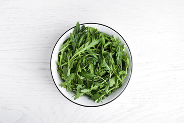 Fresh arugula in bowl on white wooden table, top view