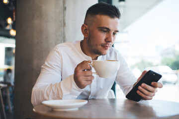 Focused man browsing smartphone while drinking coffee