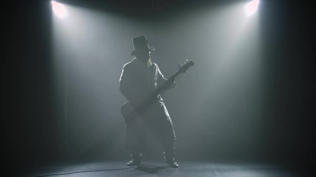Bearded rock bass guitarist plays a solo part during a studio performance with smoke and neon lights. Silhouette of a man in a hat and a long cloak plays the guitar in slow motion.