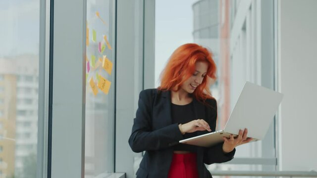 Office work with laptop and stickers. Young red-haired woman holds a laptop in her hand and takes a sticker from the window and examines it. Business lady in the office at work.