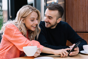 Happy couple in love having fun on leisure laughing at funny photo on smartphone sitting in cafe,cheerful young caucasian woman and man enjoying news from social networks talking using mobile phone