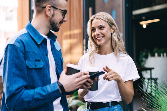 Positive caucasian woman pointing on mobile phone of her male friend colleagues discussing new apps on coffee break, 20s hipster guy holding smartphone talking to girl on free time near cafe