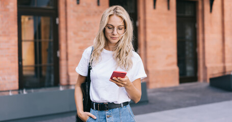 Attractive caucasian blonde female in eyewear reading income message on smartphone standing on...