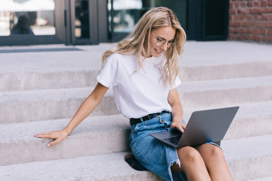 Cheerful caucasian female in eyewear for vision correction working remotely satisfied with 4G internet connection, smiling woman blogger watching video online on laptop computer sitting outdoors
