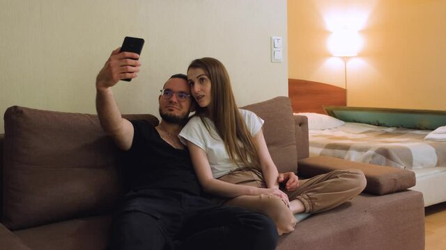 A young charismatic guy with a beard in glasses and a cute blonde with braces on her teeth make a selfie on a smartphone, sitting on the couch, hugging each other.