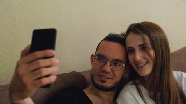 A young charismatic guy with a beard in glasses and a cute blonde with braces on her teeth make a selfie on a smartphone, sitting on the couch, hugging each other. Close-up of faces.