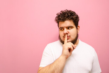 Serious guy in a white T-shirt and overweight looks aside with a pensive face and shows a sign of silence. Concentrated fat man with a beard shows the gesture "Shsh" and looks at copy space.