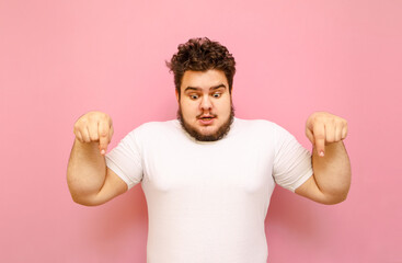 Shocked overweight guy in white t-shirt isolated on pink background, looks down and shows on copy...
