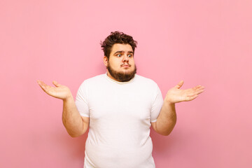 Portrait of puzzled fat man with shocked face on pink background, looking to the side and spreading...