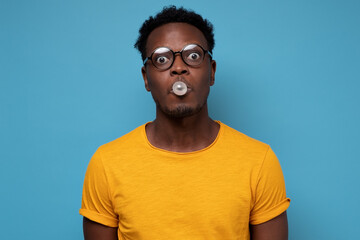Young african american man in glasses inflating ball with chewing gum while looking at camera with...